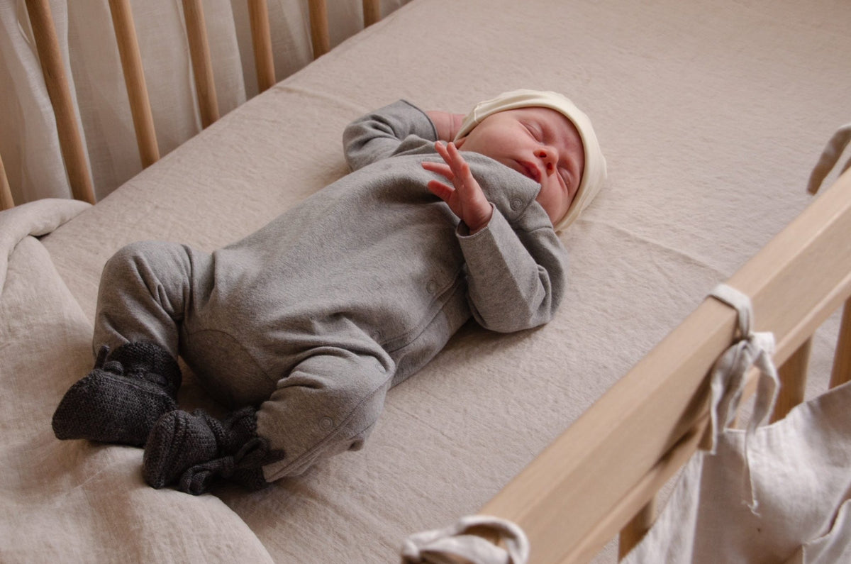 Newborn asleep in a wooden crib wearing the Gray Label Baby Suit in grey melange with a cream hat and knitted booties