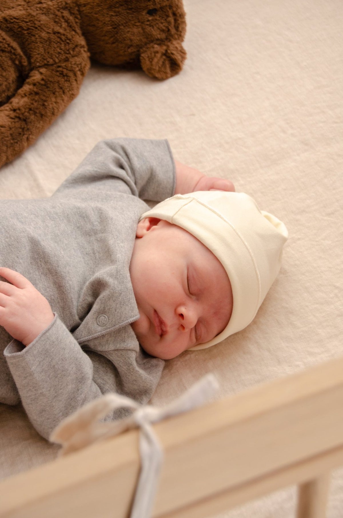 Newborn baby sleeping in a wooden crib wearing a cream hat, dressed in the Gray Label Newborn Suit in Grey Melange