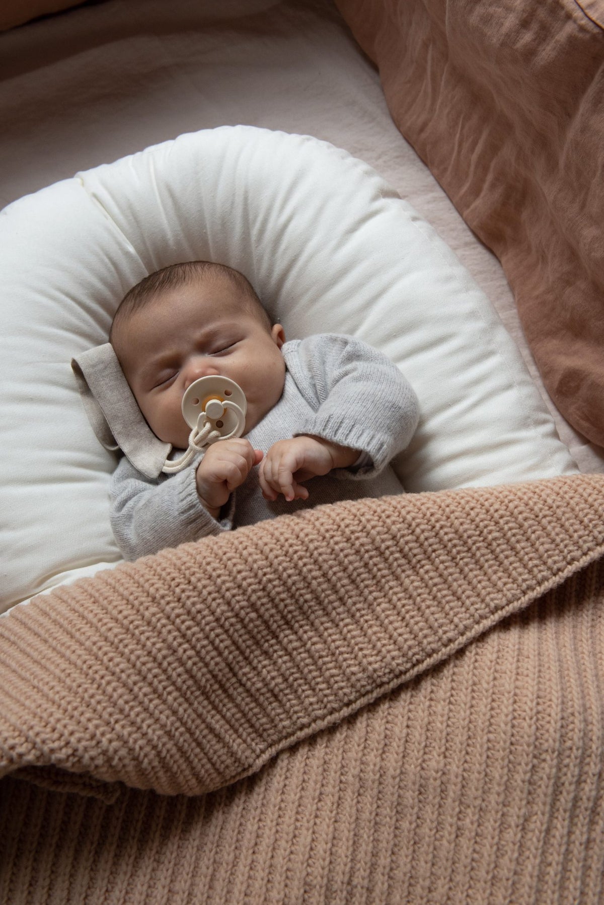 Close-up of a sleeping baby wrapped in a Hazel & George Abel Chunky Blanket in Biscuit, lying in a cushioned nest with pink linen bedding