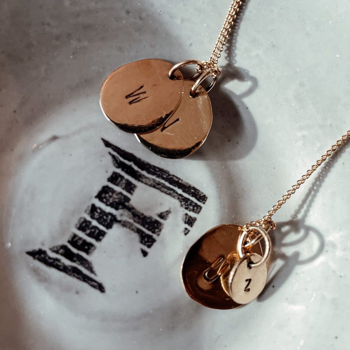 Hazel & George Initial Hanger Large 14K Gold, two gold pendants engraved with the letter M resting on a decorative plate with a fox illustration, next to a necklace with a small Z charm