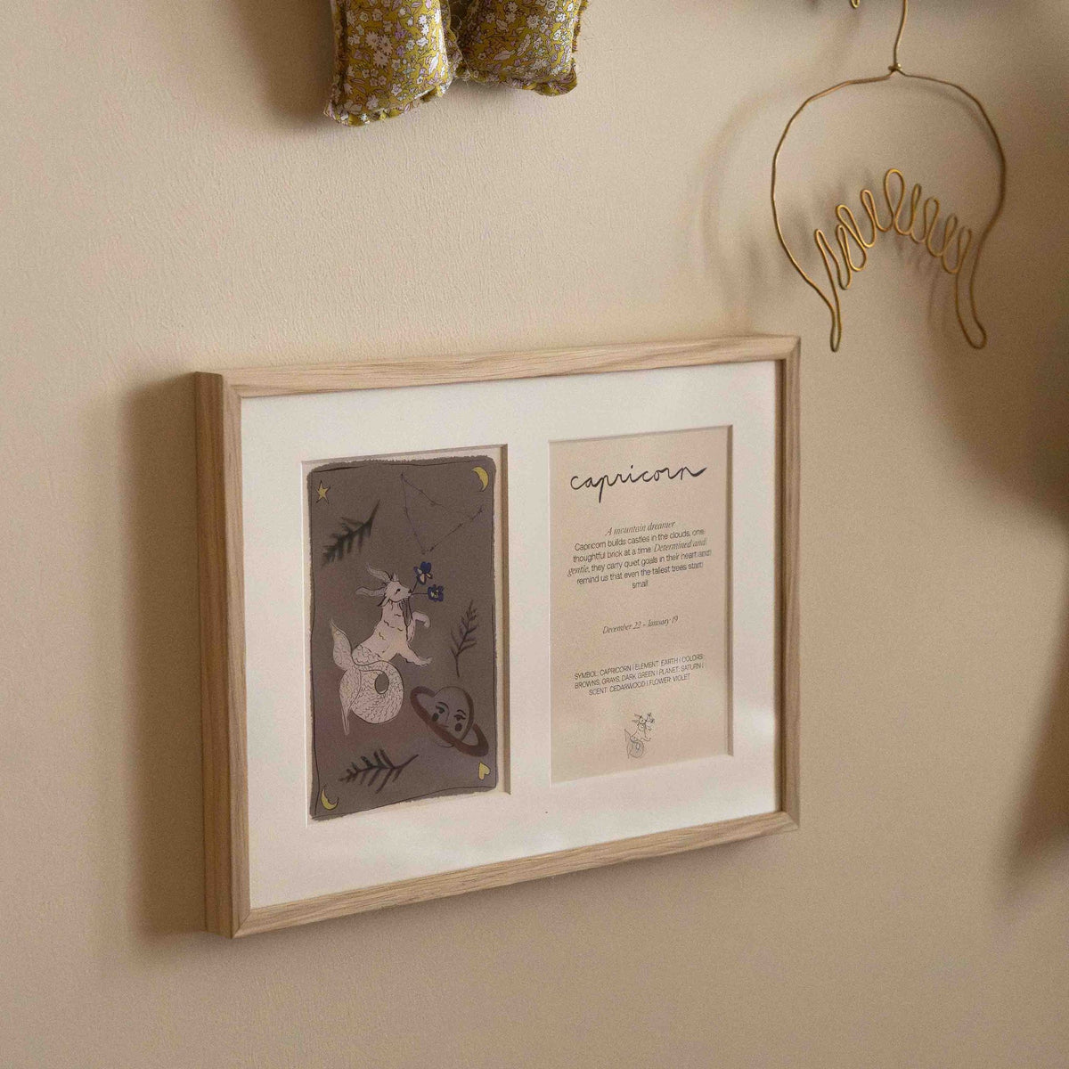 Ingelijste dubbele Hazel & George Steenbok-poster aan de wand, gezien vanuit een hoek, naast een stoffen letterkussen en een hangend kledinghangertje van metaaldraad