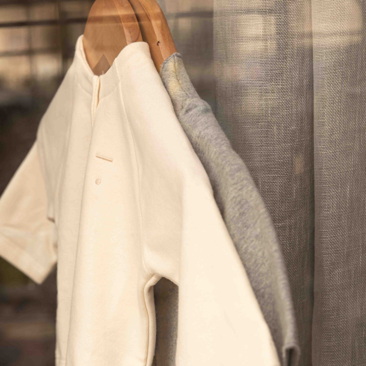 Close-up of the Gray Label Baby Cardigan in Cream, hanging next to a grey sweater near a window