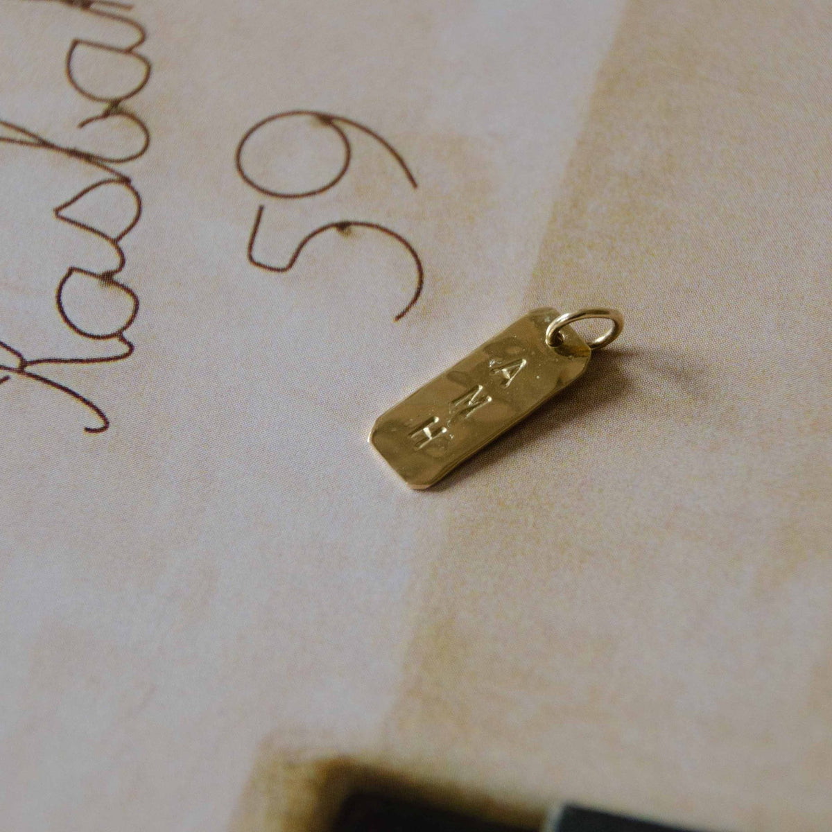 Hazel & George Rectangular Initial Hanger 14K Gold, rectangular pendant engraved with the letters AMH lying on a vintage handwritten letter