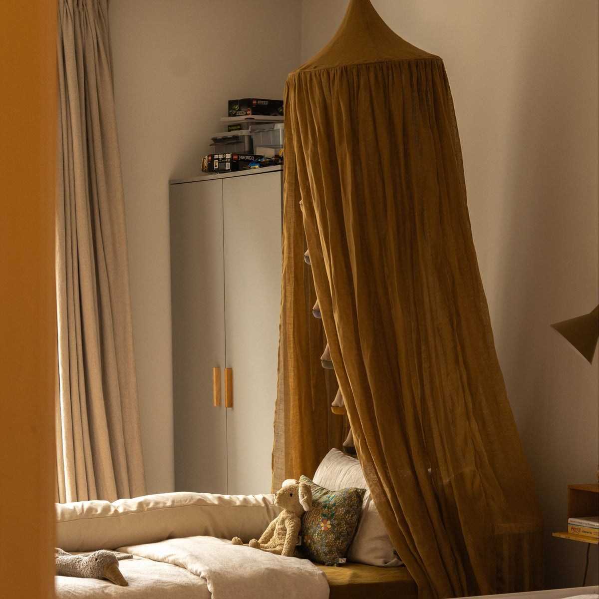 Hazel & George Linen Bed Canopy in Dore hanging above a bed with plush toys, in a bedroom with a wardrobe and toy boxes