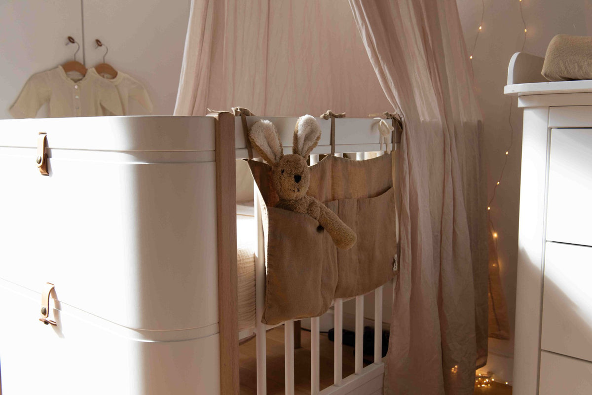 Hazel & George Linen Bed Pocket in Dore on a white cot rail, in a nursery scene with a canopy, fairy lights and a plush bunny in the pocket