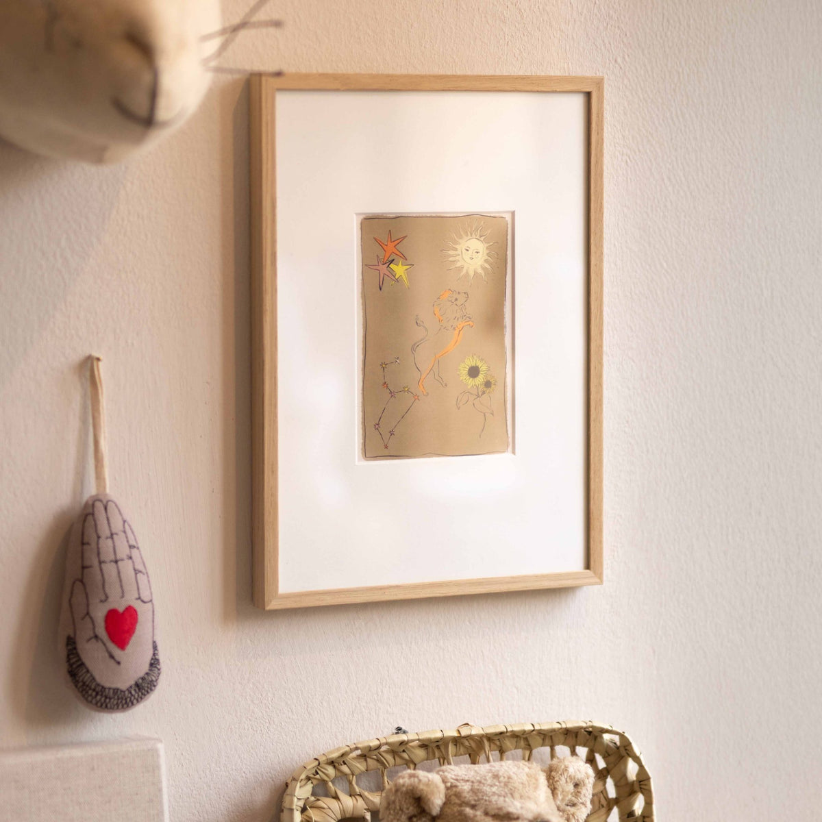 Ingelijste Hazel & George Leeuw-poster aan de wand in een kinderkamer, naast een hangende hartje-in-hand decoratie en een mandje met een teddybeer