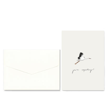 A minimalist greeting card and matching envelope set on a white background. The envelope is plain white, and the card features a small black-and-white flying stork illustration above the handwritten text &