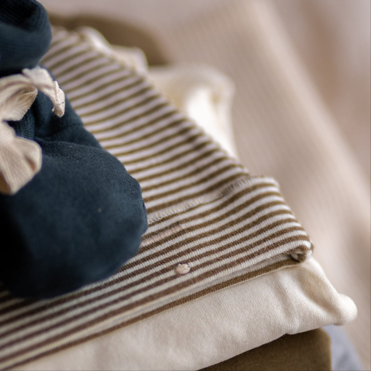 Gray Label Baby Beanie in Woody Cream, styled on top of folded clothing next to a pair of navy booties
