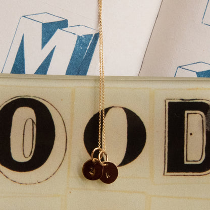 Hazel &amp; George Hangertje Met Initiaal Klein 14K Goud, gouden ketting met twee kleine hangertjes met initiaal, gefotografeerd tegen een grafische achtergrond met grote letters