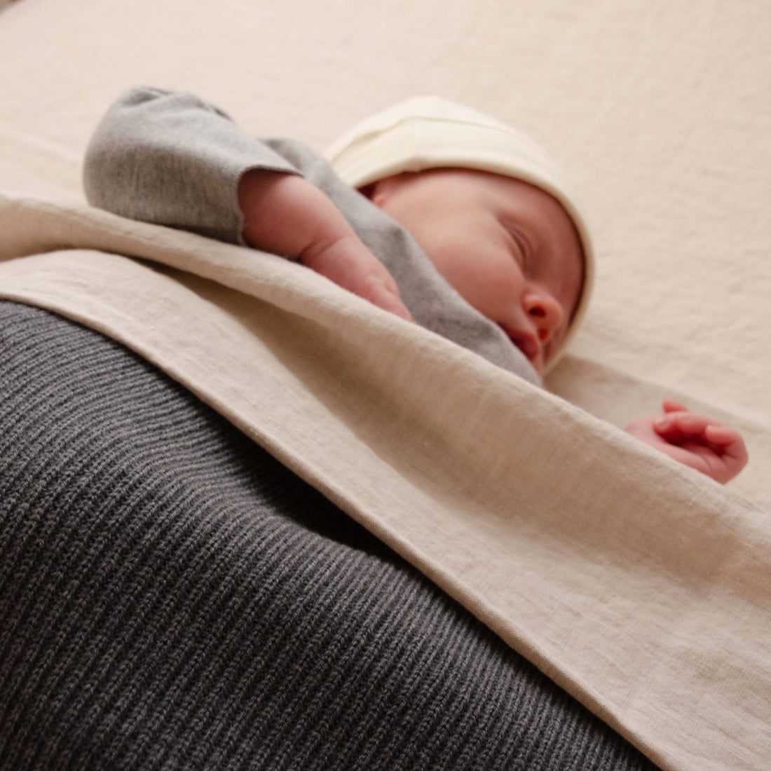 Sleeping newborn baby wearing a cream hat, covered with a Hazel &amp; George Georgia Merino Blanket in Charcoal