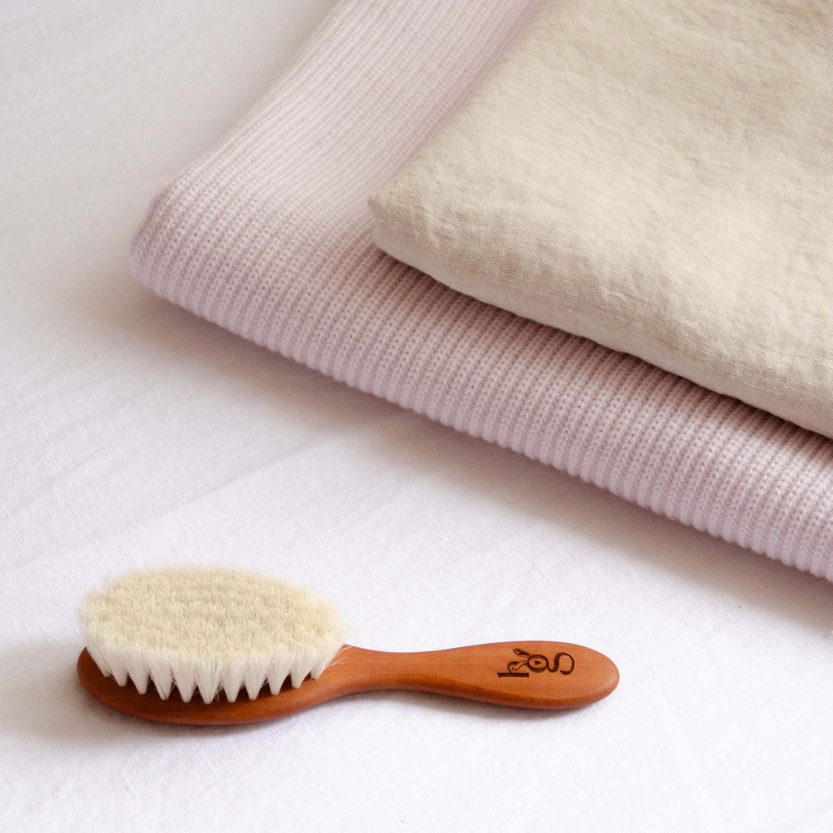 Wooden baby hair brush styled with a folded Hazel &amp; George Georgia Merino Blanket in Poudre and cream linen fabric