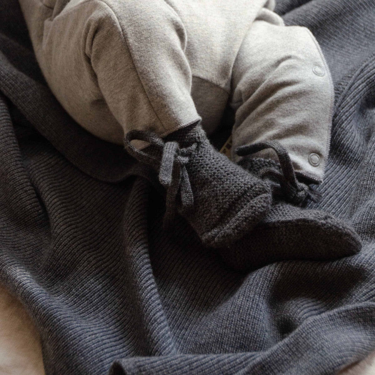 Baby wearing knitted booties, sitting on a Hazel &amp; George Hazel Merino Baby Blanket in Charcoal