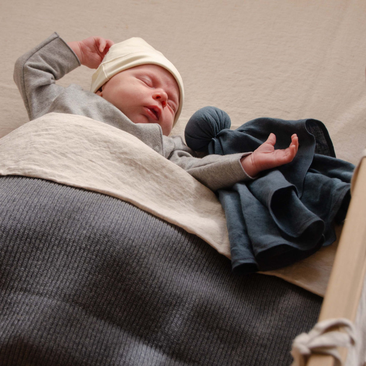 Sleeping baby wearing a cream hat in a crib, covered with a Hazel &amp; George Hazel Merino Baby Blanket in Charcoal