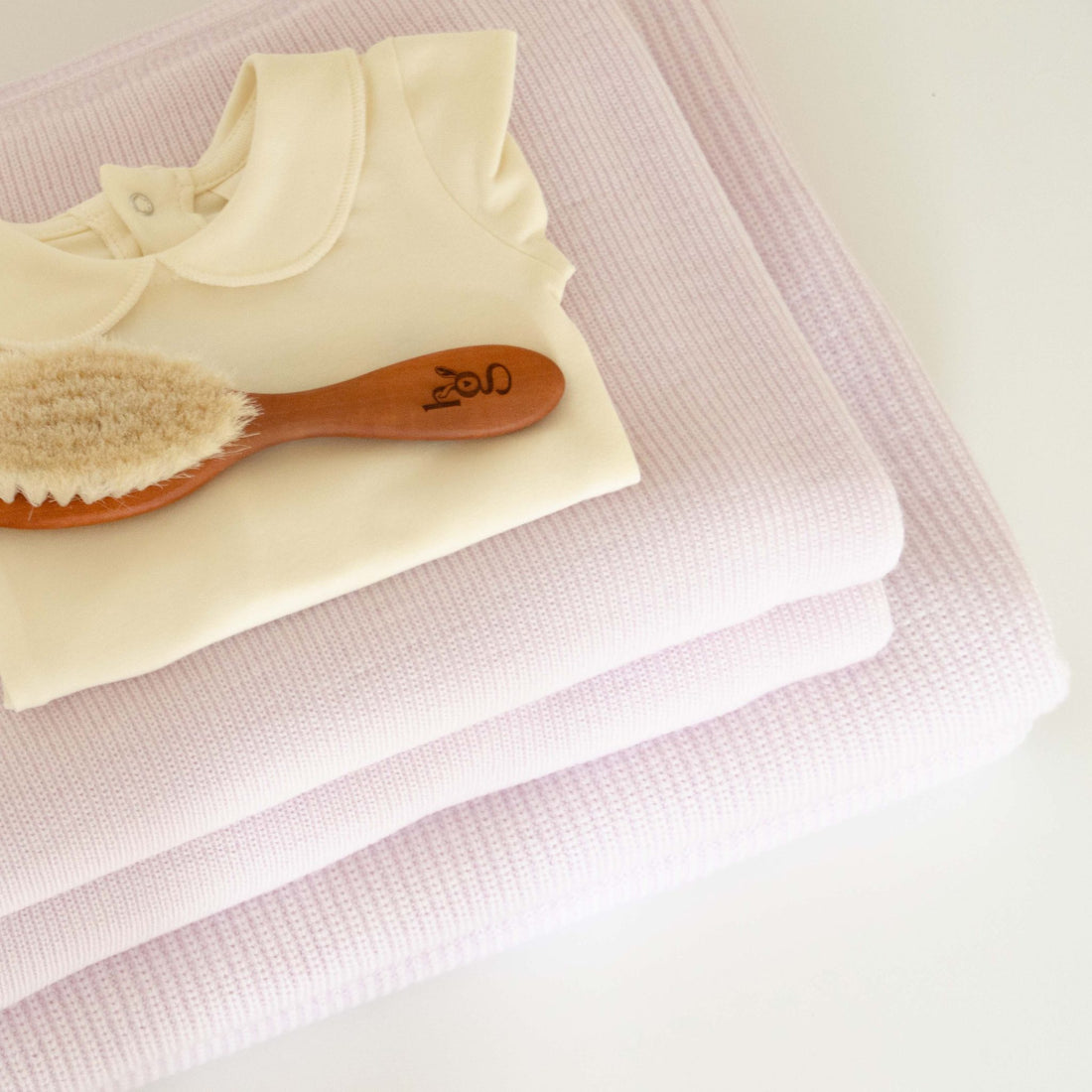 Folded Hazel &amp; George Hazel Merino Baby Blanket in Poudre, styled with a cream baby onesie and a wooden baby hair brush