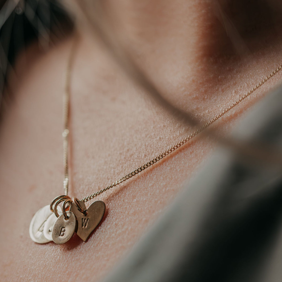Hazel &amp; George Heart Initial Hanger, close-up of a heart-shaped pendant engraved with the letter W worn on the skin, layered with small round initial charms