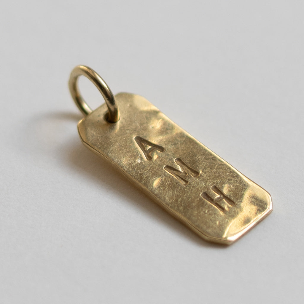 Close-up of the rectangular pendant on the Hazel &amp; George Rectangular Initial Hanger 14K Gold, engraved with the letters A, M and H, on a white background