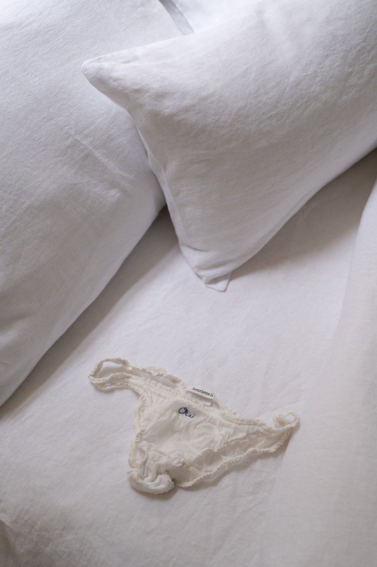 Close-up of Hazel &amp; George linen pillows in Blanc on a bed, with a small delicate baby garment on the sheet