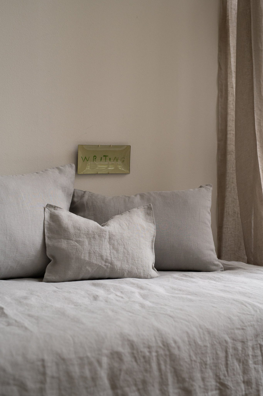 Bed styled with three Hazel &amp; George linen pillows in French Grey against a headboard, with a linen curtain nearby