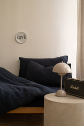 Bed with Hazel & George linen bedding in Off Black, styled bedroom corner with a nightstand lamp and speaker