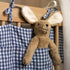 Hazel & George Linen Bed Pocket in Vichy tied to a wooden cot rail, with a plush bunny toy hanging from the pocket