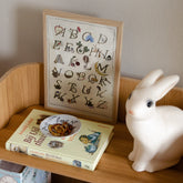 Ingelijste Hazel & George alfabetposter (limited A4-editie) op een houten plank, naast een konijnvormig nachtlampje en een kinderboek