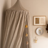 Hazel & George Linen Bed Canopy in Naturel in a nursery corner, with a star garland, framed art and a monogram plate on the wall