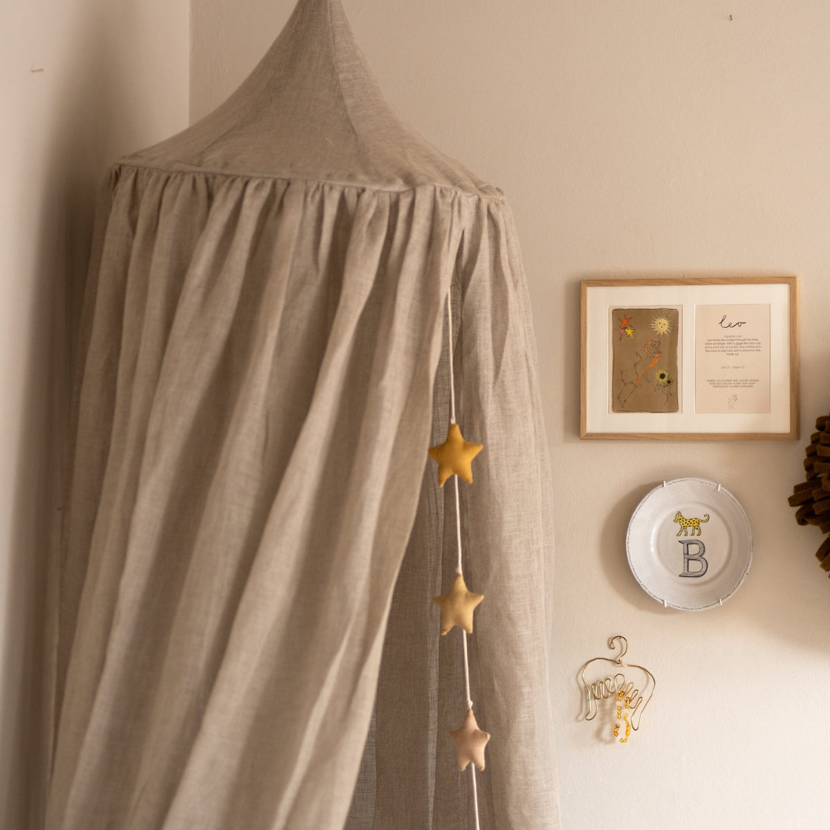Hazel &amp; George Linen Bed Canopy in Naturel in a nursery corner, with a star garland, framed art and a monogram plate on the wall