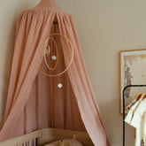 Hazel & George Linen Bed Canopy in Misty Rose hanging above a wooden cot, with a hanging mobile, framed art and a clothing rail nearby