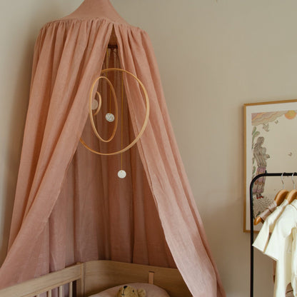 Hazel &amp; George Linen Bed Canopy in Misty Rose hanging above a wooden cot, with a hanging mobile, framed art and a clothing rail nearby