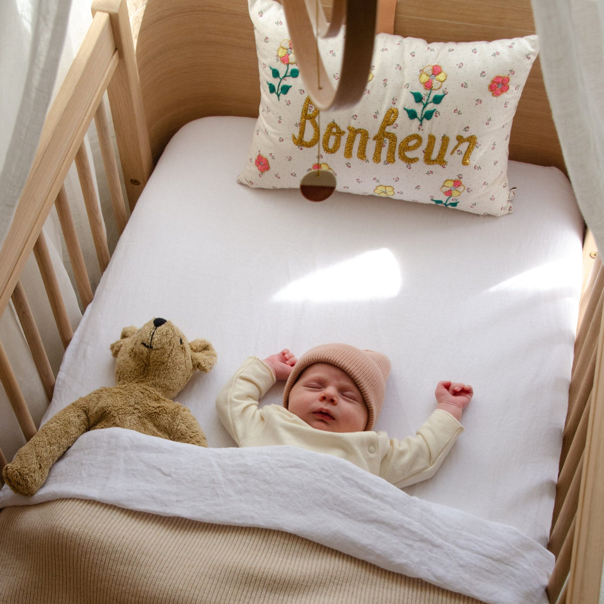 Hazel &amp; George Linen Fitted Sheet in Blanc on a cot, with a sleeping baby, a teddy bear and a decorative cushion