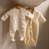 The Gray Label Newborn Suit in Cream hanging on a wooden hanger next to a knitted cream sweater vest