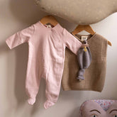 The Gray Label Newborn Suit in Faded Pink hanging on a wooden hanger next to a knitted vest and a soft fish toy