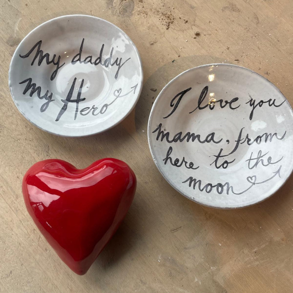 A red porcelain heart-shaped decoration accompanied by two white dishes with black scripted text on them, placed on a neutral background.