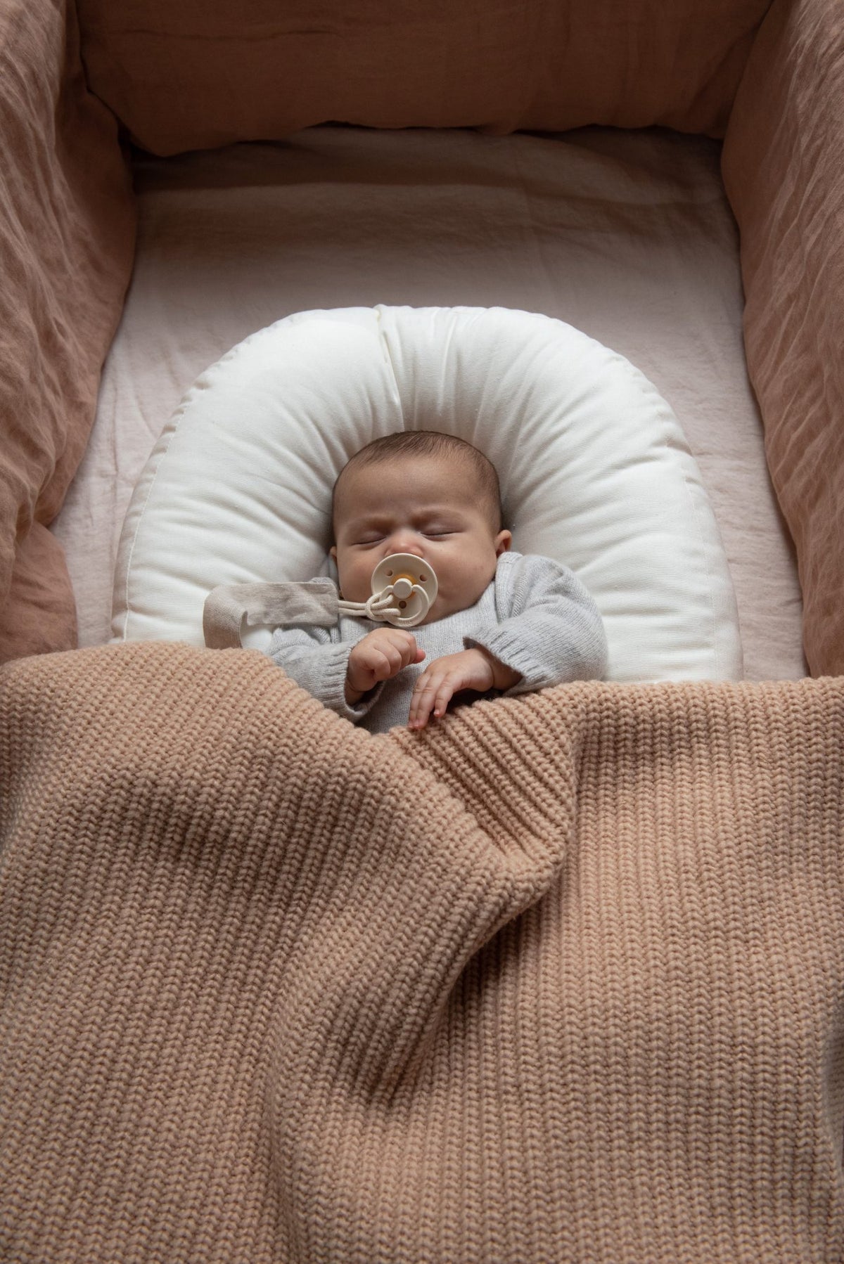 Sleeping baby in a crib with pink linen bedding, covered with a Hazel &amp; George Abel Chunky Blanket in Biscuit