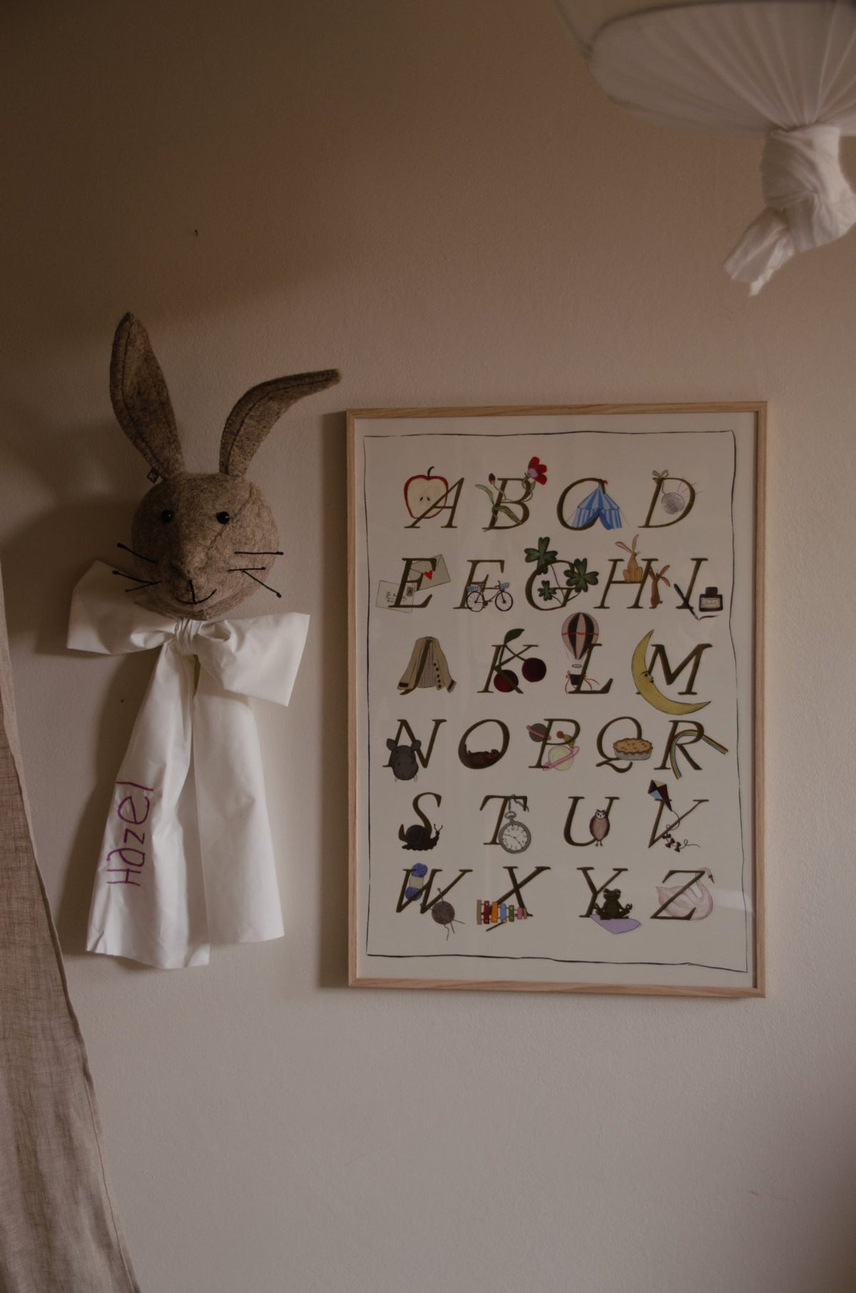 Ingelijste Hazel &amp; George alfabetposter aan de wand in een kinderkamer, naast een knuffelkonijnenkop met strik