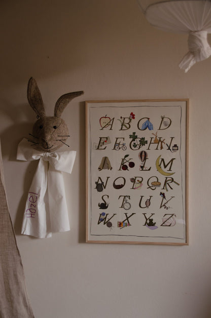 Ingelijste Hazel &amp; George alfabetposter aan de wand in een kinderkamer, naast een knuffelkonijnenkop met strik