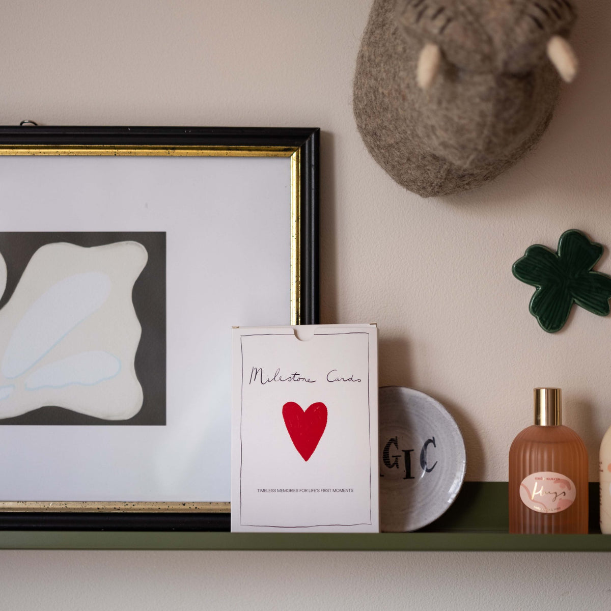 Milestone Cards box displayed on a shelf with framed art, a felt animal head, a ceramic clover and perfume bottles
