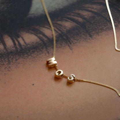 Hazel &amp; George Chunky Initiaal 14K Goud, gouden ketting met chunky letterkralen die MOSS vormen, gefotografeerd op een artistieke geprinte achtergrond