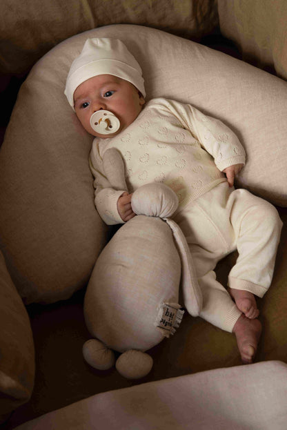 Baby lying in a Hazel &amp; George linen nursing pillow in Naturel, dressed in a cream knit outfit and holding a plush toy