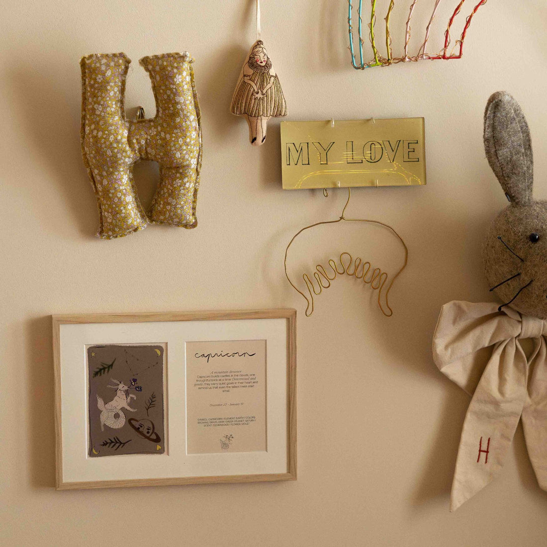 Ingelijste dubbele Hazel &amp; George Steenbok-poster aan de wand tussen kinderkamerdecoratie, met een gevlochten letter H-kussen, een pluche konijn en een gouden &