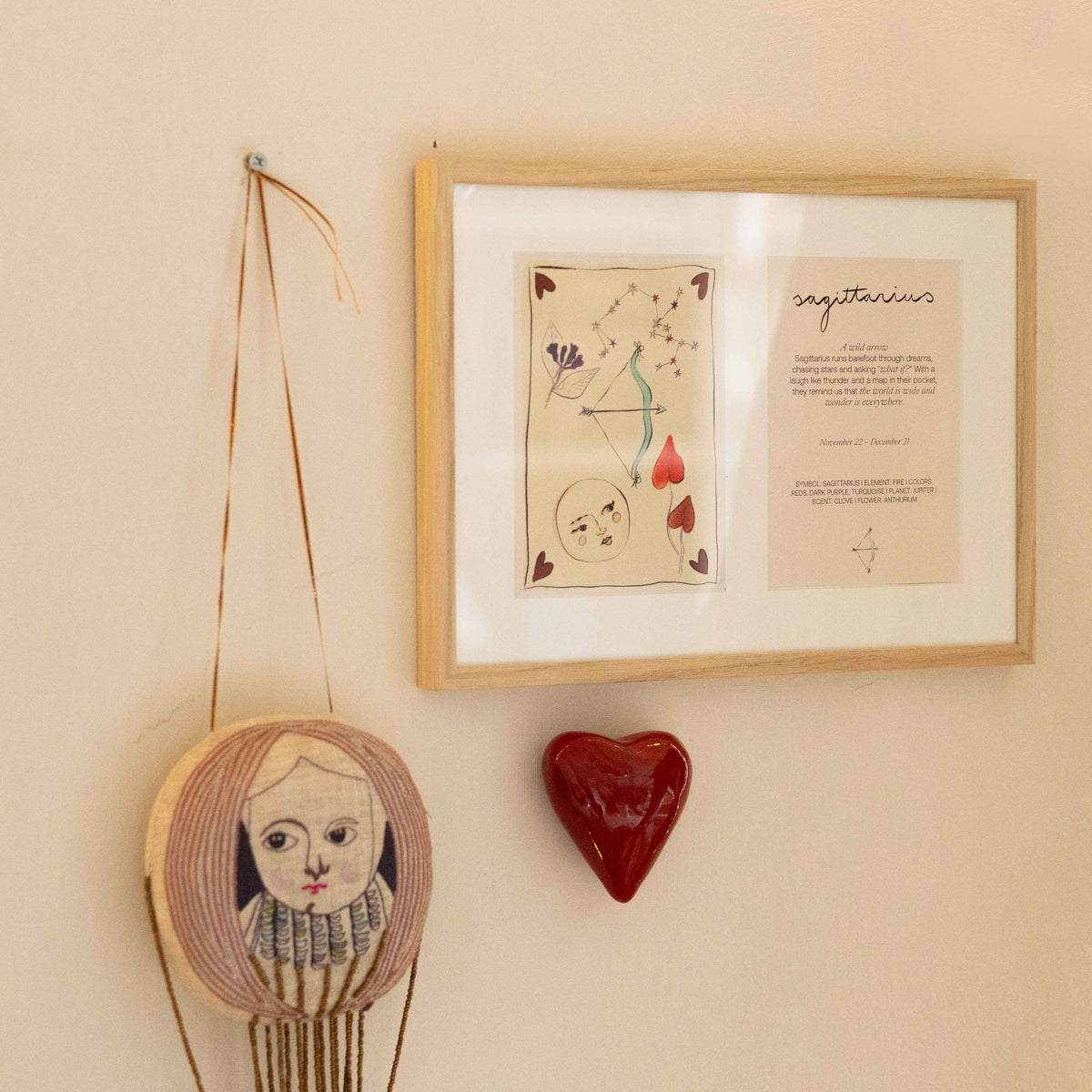 Ingelijste dubbele Hazel &amp; George Boogschutter-poster aan de wand, naast een decoratieve hanger met een gezicht en een rood keramisch hartje