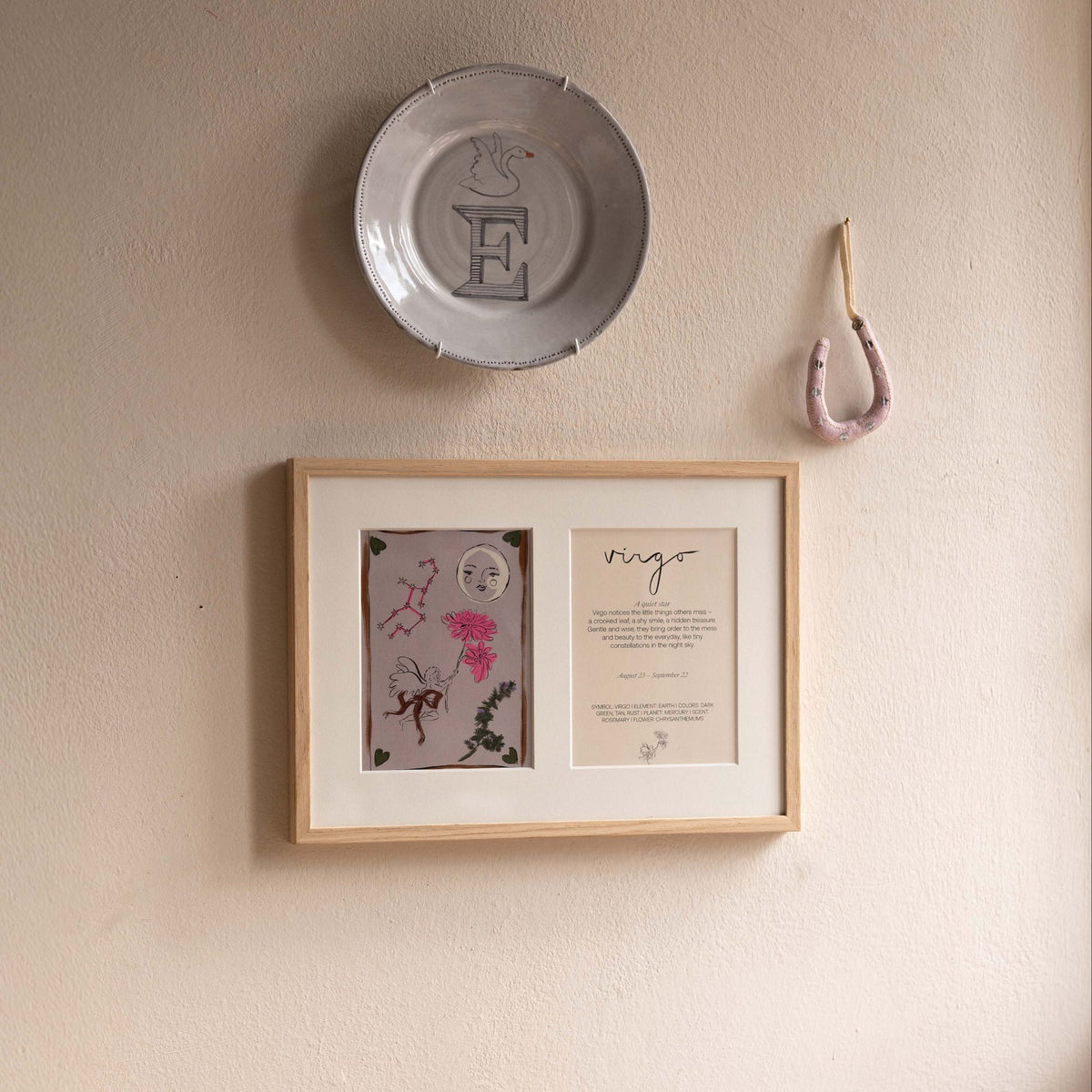 Ingelijste dubbele Hazel &amp; George Maagd-poster aan de wand, naast een sierbordje met een zwaan en de letter E en een hangend hoefijzer