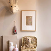 Ingelijste Hazel & George Leeuw-poster aan de wand in een kinderkamer, in een lichte houten lijst, naast een knuffelkonijnenkop en een mandje met een teddybeer