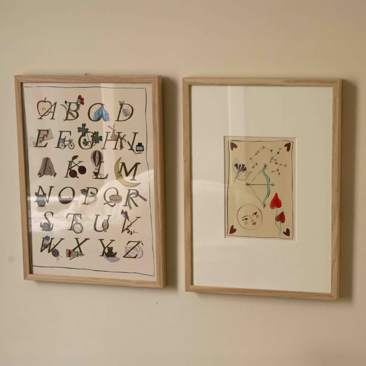Twee ingelijste Hazel &amp; George posters naast elkaar aan de wand: een alfabetposter en een Boogschutter-poster