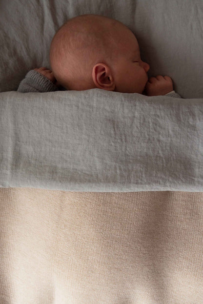 Close-up of a sleeping baby resting on linen bedding, tucked under a Hazel &amp; George Georgia Merino Blanket in Milk