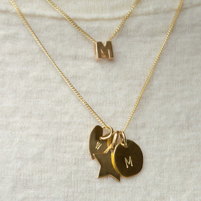 Hazel &amp; George Star Initial Hanger, layered gold necklaces with a chunky letter M, a star-shaped pendant, and a round pendant engraved with the letter M, on a knit sweater