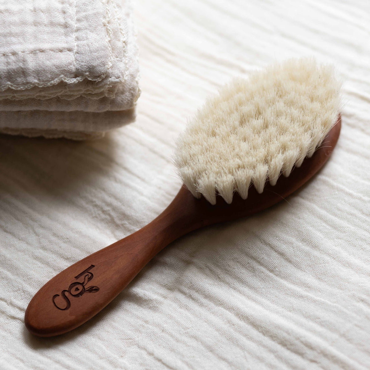 Hazel &amp; George wooden baby hair brush lying on folded muslin bedding next to a muslin blanket