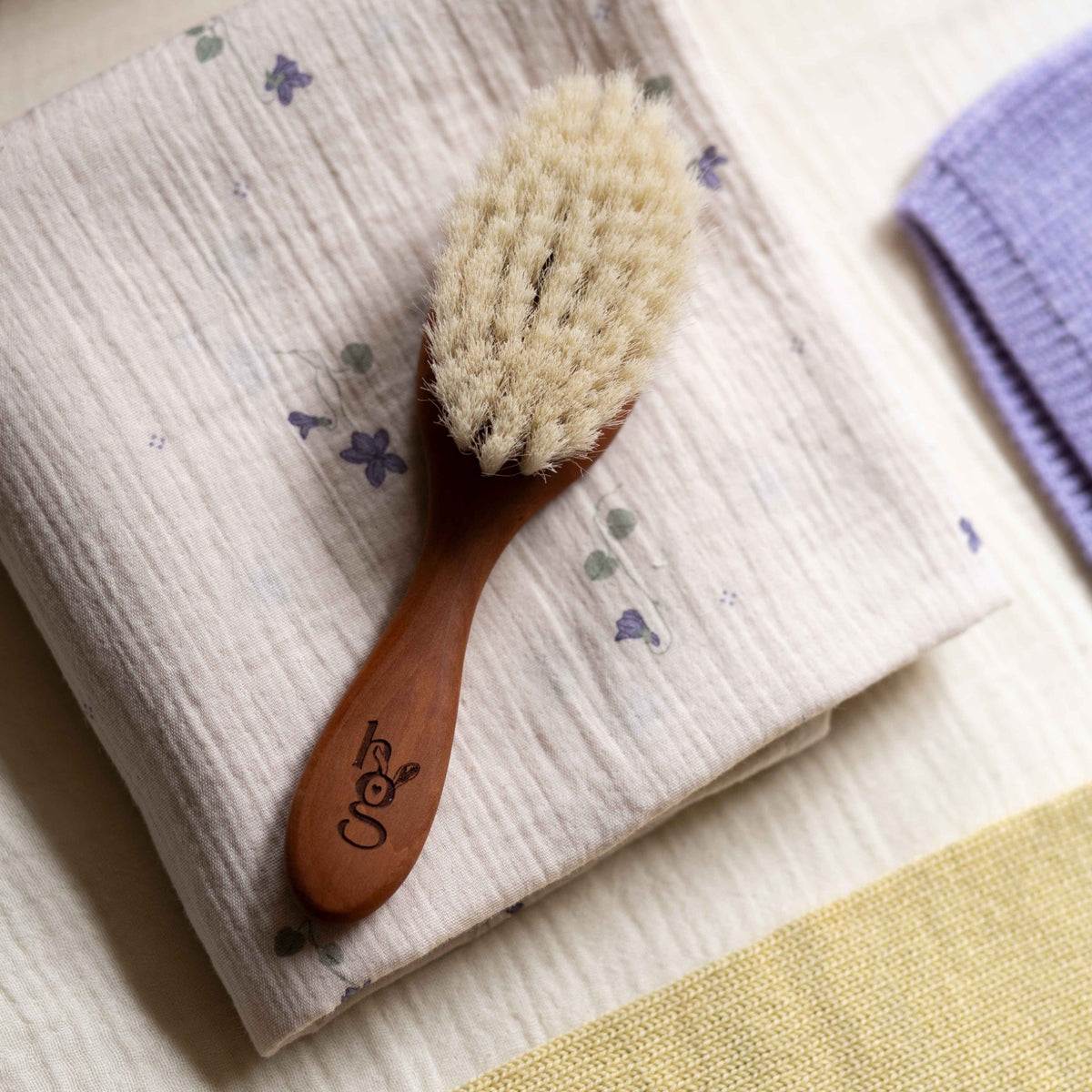 Hazel &amp; George wooden baby hair brush lying on a violet-print muslin swaddle blanket