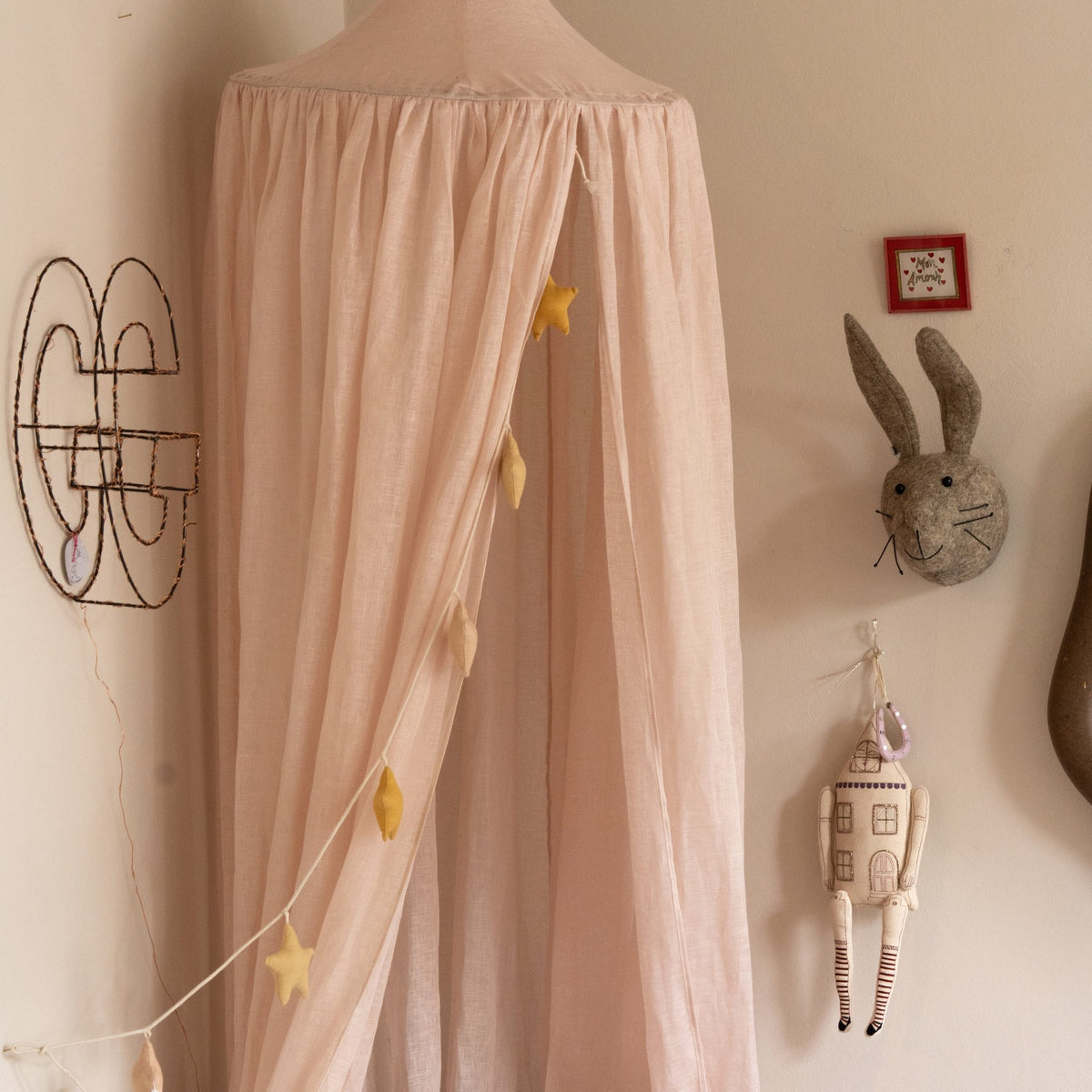 Hazel &amp; George Linen Bed Canopy in Craie in a nursery, with a star garland hanging inside, a felt rabbit head and decorative wall pieces