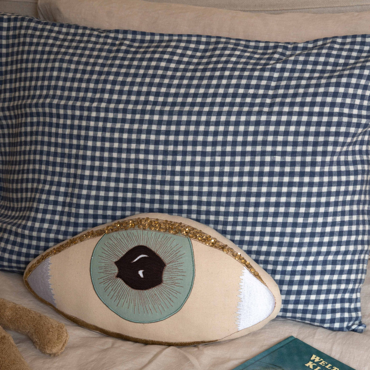 Hazel &amp; George linen pillow in Vichy styled on a bed with a decorative eye-shaped pillow, a plush toy and a book